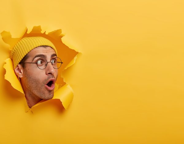 Stunned man looks with great surprisement or fear aside, opens mouth widely, wears hat and round glasses, looks through paper hole, isolated over yellow background. Shocking relevation concept