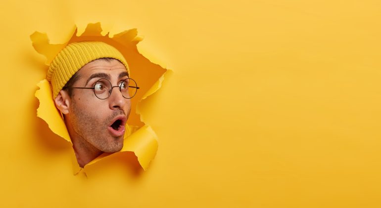 Stunned man looks with great surprisement or fear aside, opens mouth widely, wears hat and round glasses, looks through paper hole, isolated over yellow background. Shocking relevation concept Stunned man looks with great surprisement or fear aside, opens mouth widely, wears hat and round glasses, looks through paper hole, isolated over yellow background. Shocking relevation concept