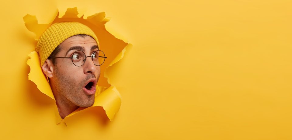 Stunned man looks with great surprisement or fear aside, opens mouth widely, wears hat and round glasses, looks through paper hole, isolated over yellow background. Shocking relevation concept